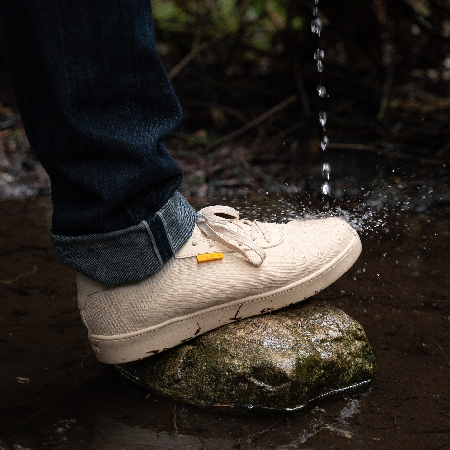 White sneaker stepping onto a rock with water droplets falling, surrounded by natural elements.